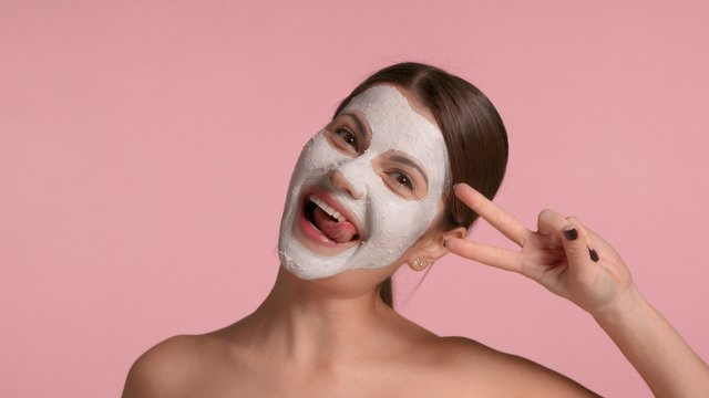 30s brunette woman with a facial clay mask on laughing and having fun making faces in studio on pink background