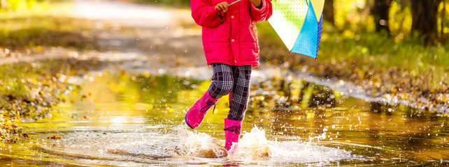 Very cute little girl in pink Jacket and rubber boots is jumping over a puddle on a rainy day