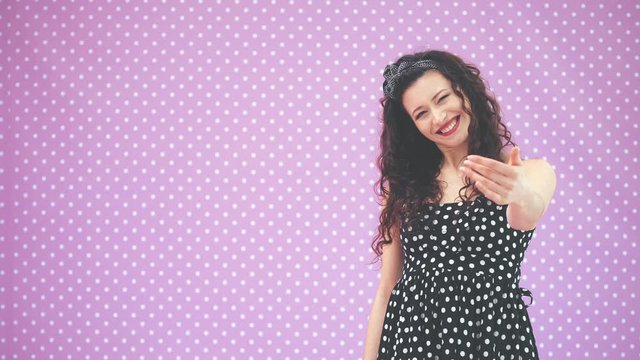Lovely young girl with kinky hair, in black polka-dots dress dancing, waving her hands, calling to join her, pointing at copy space for text or product.