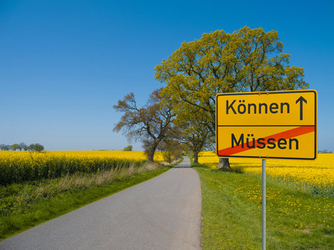 landschaft mit stra&szlig;e furch ein rapsfeld und einem Ortsschild mit den Worten K&ouml;nnen und M&uuml;ssen