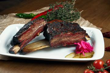 Close up view on delicious appetizing smoked and grilled beef ribs. Barbecue ribs - a classic dish of American cuisine on a white plate. Wooden background