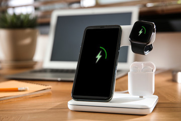 Mobile phone, earphones and smartwatch charging with wireless pad on wooden desk. Modern workplace...