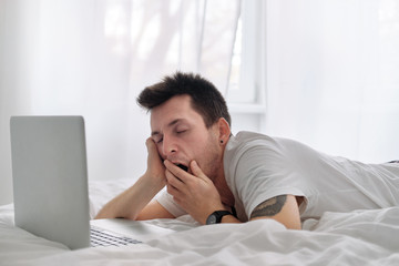 Young man in the white shirt on a bed with white linen is upset looking at the screen of a laptop.