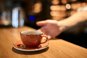 Close up cropped photo of man serving coffee while standing in coffee shop. Focus on orange cup of tea, blurred background.