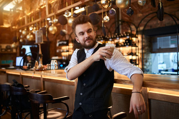 happy young man holding a glass of latte, gets pleasure from tasting it, leaning on the table and looking aside, guy having a rest at work