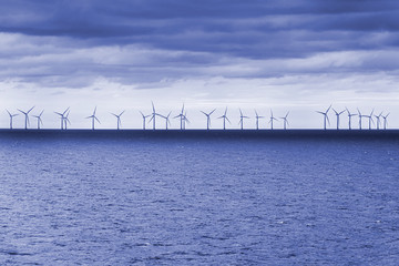 Wind farm with big offshore windmills at the horizon, calm ocean sea water and cloudy sky in background in blue coloration - concept power generation climate change business electrical energy nature