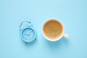 Cup of morning coffee and alarm clock on light blue background, flat lay