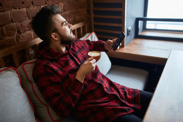 young thoughtful pensive man looking at the window, holding a smart phone and cup of tasty tea, thinking about business, life, woman