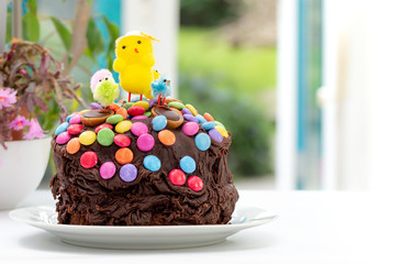 Fun kids chocolate Easter cake decorated by a child with chocolate frosting covered in colorful chocolate beans and Easter chicks. Decoration sliding down sides of cake. In front of garden window.