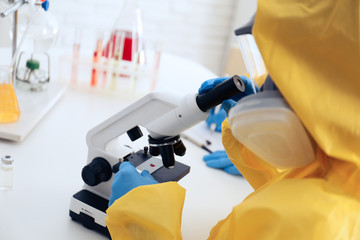 Scientist in chemical protective suit using microscope at table, closeup. Virus research