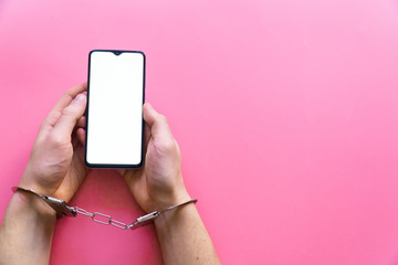 Men's hands in handcuffs hold a smartphone on a pink background. The concept of internet and gadget dependency.