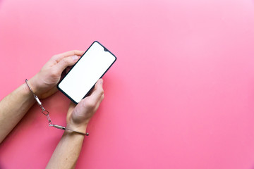 Men's hands in handcuffs hold a modern smartphone with blank screen on pink background. The concept of internet and gadget dependency.
