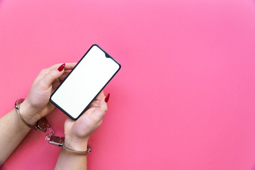 Woman hands in handcuffs hold a smartphone on a pink background. The concept of internet and gadget dependency.