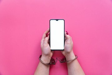 Woman hands in handcuffs hold a smartphone with blank screen on a pink background. The concept of internet and gadget dependency.