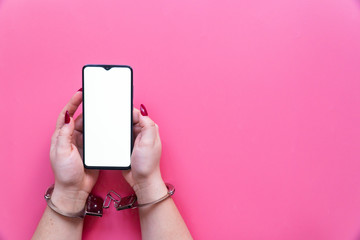 Woman hands in handcuffs hold a modern smartphone with blank screen on a pink background. The concept of internet and gadget dependency.