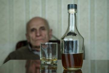 blurry old man looks at bottle and glass with cognac on brown wooden table at home