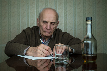 old man writes testament with pen sitting at brown wooden table by whiskey bottle
