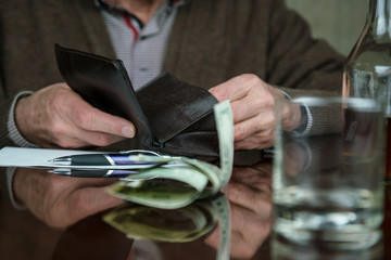 aged man hands on black leather wallet on brown wooden table with banknotes and alcohol