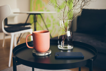 Cozy interior details. Little round table with ceramic red color cup of delicious earl gray tea. Resting after work day, offline.  Selective focus, natural light