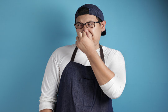 Portrait of funny young male Asian chef or waiter closing his nose, smells something bad