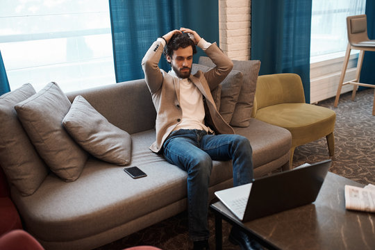 handsome bearded guy in jacket , jeans and roll neck relaxing on the couch, side view photo. man having online workshop, learning , watching vebinar, education