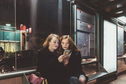 Two young beautiful women sitting bus stop using smartphone