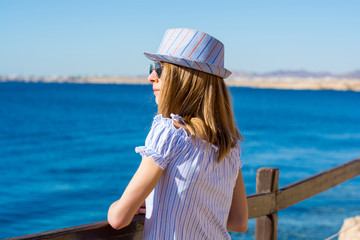 Young woman in a hat and sunglasses stands a back near the sea.
