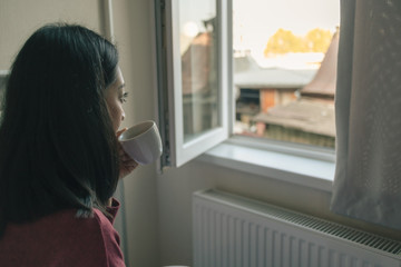 Fototapeta na wymiar Woman is drinking hot coffee and looking out to the window in her apartment.
