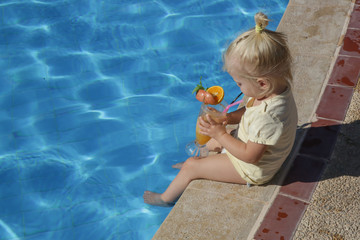 Cute blonde toddler girl sitting at the pool edge and sipping fresh squizeed orange juice, a day at the swimming pool, hydratation and vitamin intake during hot summer days