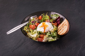 Healthy and Tasty breakfast food: green salad with poached egg, cherry tomatoes and peace of bread in round plate; fork and knife, saltcellar and breadbin near the plate on  dark grey background. 