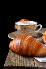 Golden croissants of yeast dough and a cup of coffee on the edge of the wooden table against black background