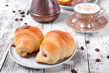 Two croissants of yeast dough on ceramic plate and freshly brewed coffee in a cup on white wooden table. Breakfast idea