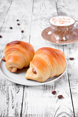 Two croissants of yeast dough on ceramic plate and freshly brewed coffee in a cup on white wooden table. Breakfast idea