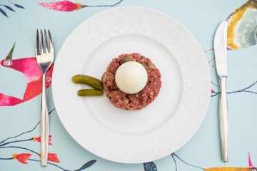 A Raw steak tatar with a boiled egg and gurkins on a white plate with cuttlery on a decorative set table