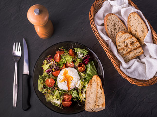 Healthy and Tasty breakfast food: green salad with poached egg, cherry tomatoes and peace of bread in round plate; fork and knife, saltcellar and breadbin near the plate on  dark grey background. 