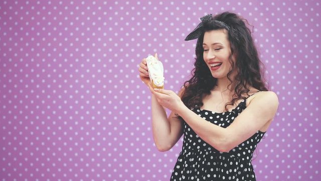 Lovely young curly girl standing, showing to the camera creamy custard tart with pink topping and marshmallow, smelling it, smiling happily.