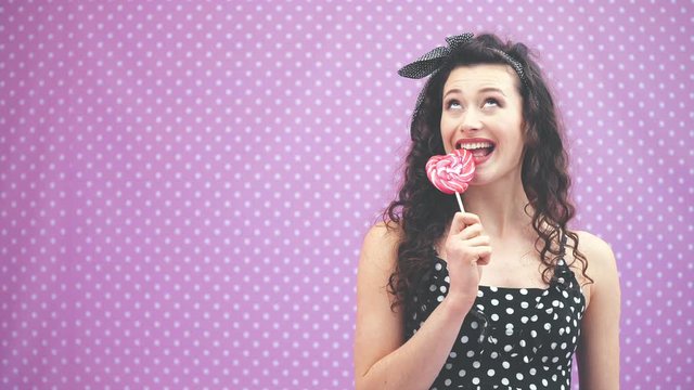 Lovely young curly girl standing, holding sweet red lollipop on the stick in her hand, smiling joyfully.