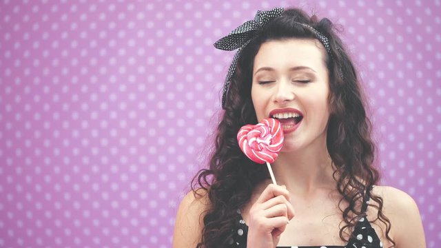Lovely young curly girl trying to bite a piece of sweet red lollipop on the stick, but it is too hard.