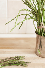 A bunch of wild asparagus lies on a wooden top and another bunch into a metal pot, with a worm crawling up on it, in a home kitchen.