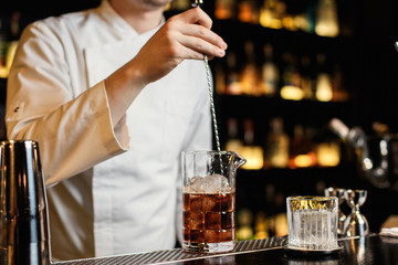 A horizontal image of a rocks glass with ice and bartender stirring a cocktail in a mixing glass...