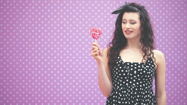 Lovely young curly girl standing, holding sweet red lollipop on the stick in her hand, petting her belly, looking at the candy, showing tisk gesture.
