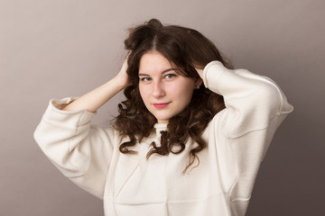 Naklejka premium Beautiful young brunette teenager girl with wavy long curly thick hair in a white sweater on a gray background. Waist photo