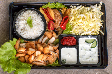 Traditional doner kebab in a black plastic container, sour cream, white boiled rice, roast meat, chiken, sauces, salad, cabbage and roasted vegetables on grey background