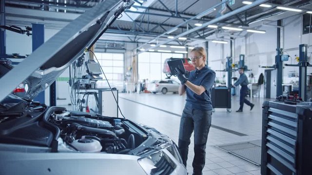 Female Mechanic Uses a Tablet Computer with an Augmented Reality Diagnostics Software. Specialist Inspecting the Car in Order to Find Broken Components Inside the Engine Bay. Modern Car Service.