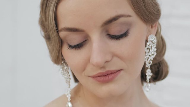 Big close up shot of young blonde bride with wedding hairstyle in tiara earrings wedding gown posing in trendy wedding salon showing gorgeous wedding makeup teasing and seductively looking in camera
