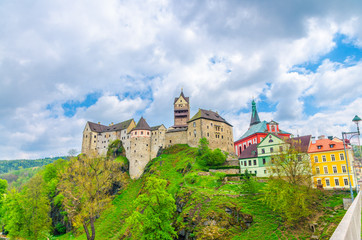 Fototapeta na wymiar Loket Castle Hrad Loket gothic style building on massive rock and colorful buildings in Loket town, green trees and hills, blue cloudy sky background, Karlovy Vary Region, West Bohemia, Czech Republic