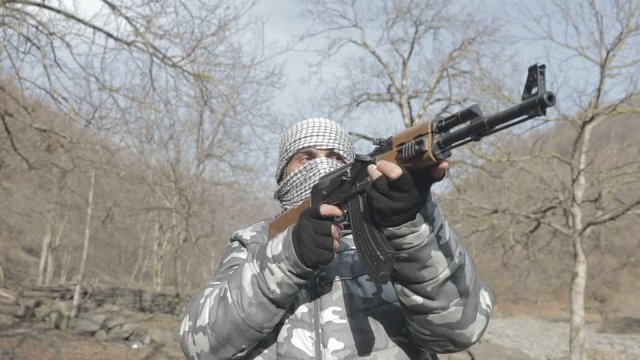 Man in mask with assault rifle ready to attack in the forest. Dangerous bandit aiming gun. Special soldier during the military operation on mountain road. Selective focus