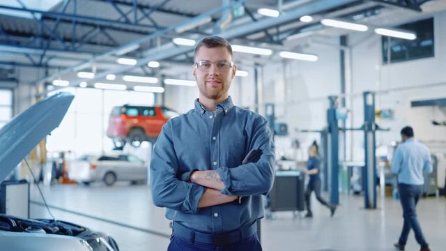 Handsome Car Mechanic is Posing in a Car Service. He Wears a Jeans Shirt and Safety Glasses. His Arms are Crossed. Specialist Looks at a Camera and Smiles.