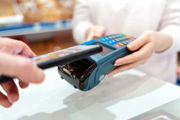 A woman's hands are held by a payment terminal and a man pays for a purchase using a smartphone. The concept of NFC, business and banking transactions