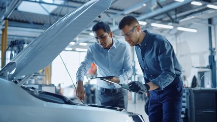 Manager Checks Data on a Tablet and Explains an Engine Breakdown to a Handsome Professional Mechanic. Car Service Employees Inspect a Car with Internal Combustion Engine. Modern Clean Workshop.